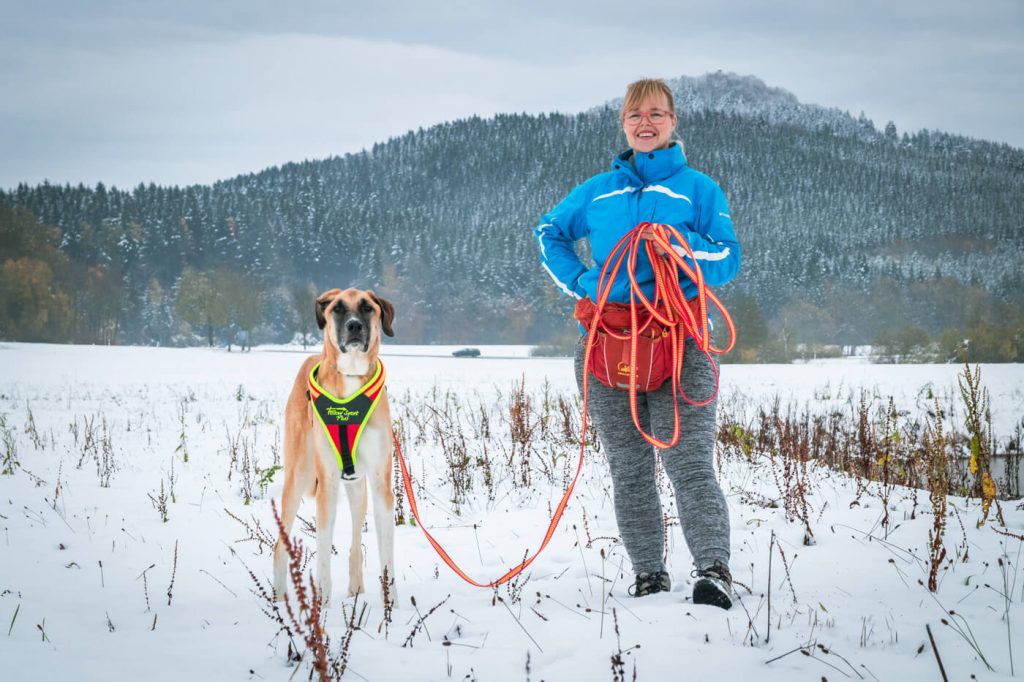 Frau posiert mit Ihrem Hund im Winter vor der Kamera mit einer Schleppleine in der Hand nach der Fährtensuche.