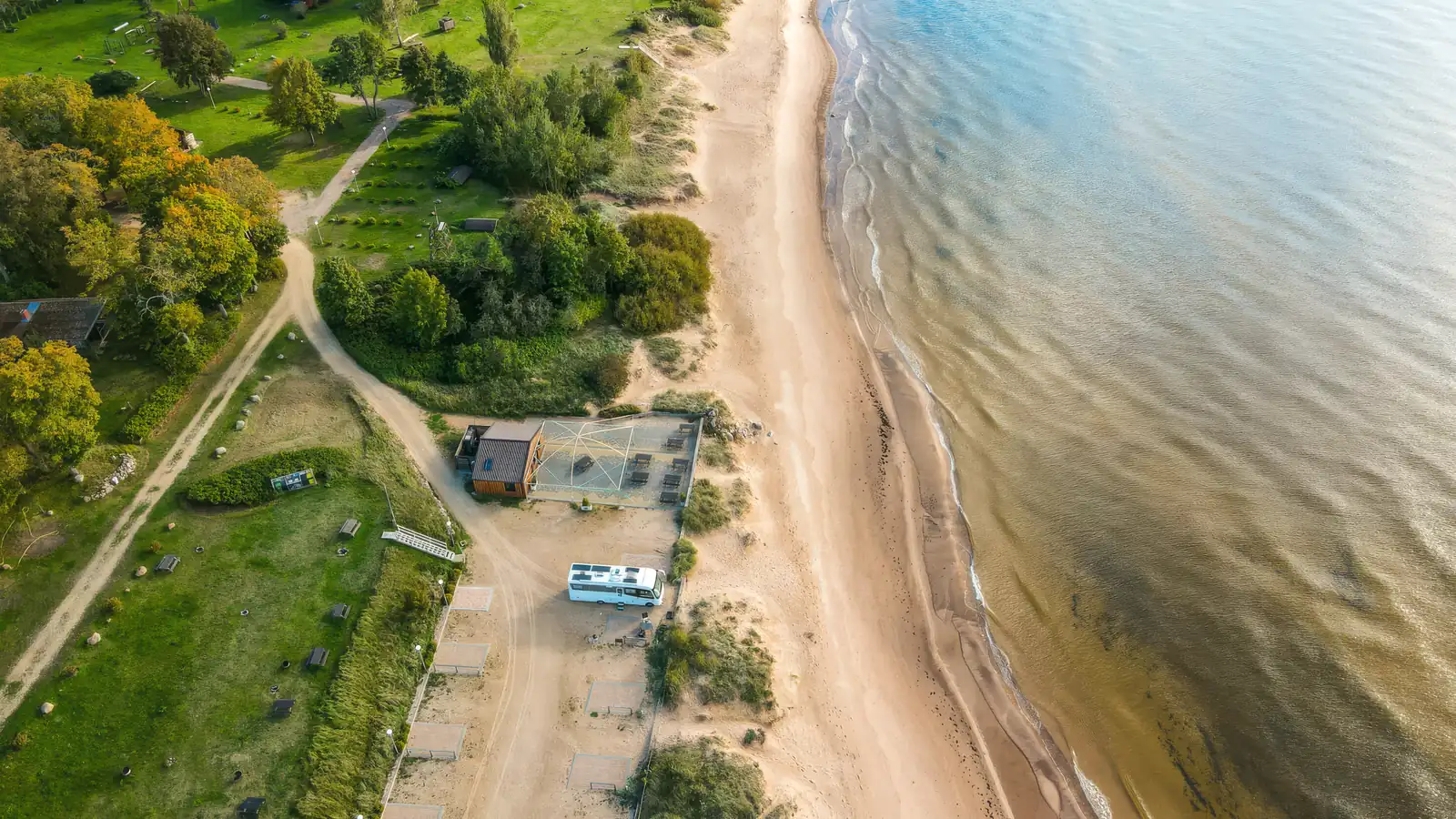 Wohnmobil auf einem Stellplatz direkt am Strand, Luftaufnahme mit Wiese, Waldweg und flach auslaufendem Sandstrand